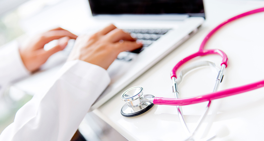 stock photo of a doctor's hands typing on a laptop with a pink stethoscope on the table